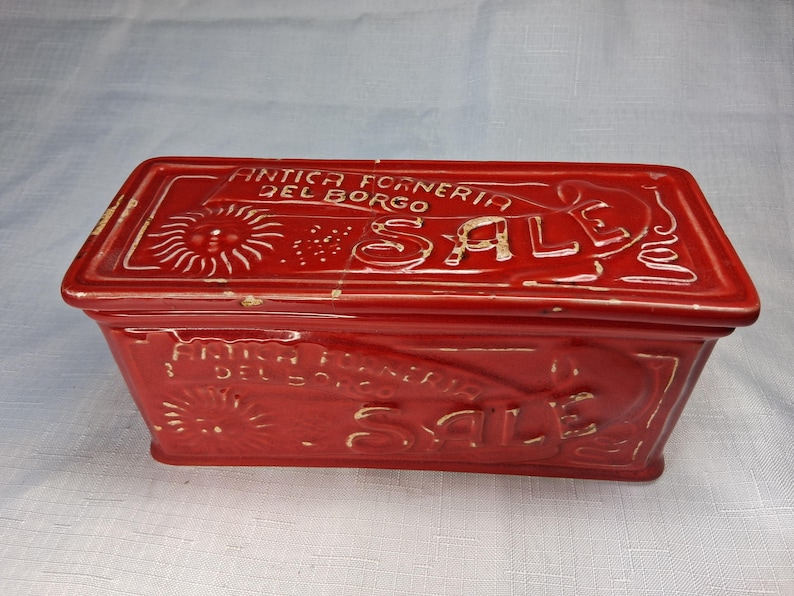 Puede incluir: Una caja rectangular de cer&aacute;mica roja con tapa. La caja presenta texto blanco en relieve que dice "ANTICA FORNERIA DEL BORGO SALE" y un dise&ntilde;o de sol. La caja es probablemente para almacenamiento o exhibici&oacute;n.