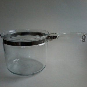 Puede incluir: Un tarro de vidrio transparente con asa y una banda metálica alrededor de la parte superior. El asa está sujeta con herrajes metálicos. El tarro es cilíndrico y parece vacío. El fondo es blanco liso.