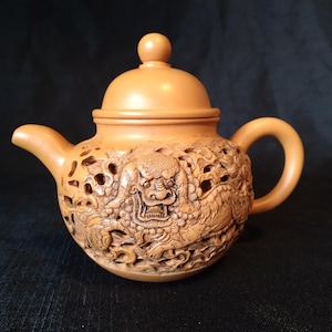 Op de afbeelding: Een bruin-beige theepot met ingewikkelde gravures van een draak en andere ontwerpen. De theepot heeft een deksel met een kleine knop, een gebogen tuit en een afgeronde handgreep. De gedetailleerde gravures zijn zichtbaar.