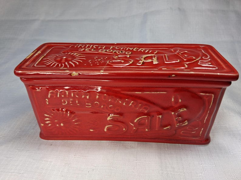 Puede incluir: Una caja rectangular de cer&aacute;mica roja con tapa. La caja tiene texto y dise&ntilde;os en relieve, incluyendo la palabra "SALE" y la frase "ANTICA FORNERIA DEL BORGO". La caja es probablemente vintage o antigua.