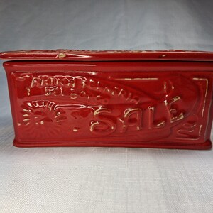 Puede incluir: Una caja rectangular de cer&aacute;mica roja con tapa. La caja presenta texto y dise&ntilde;os en relieve, incluyendo la palabra "SALE" y otro texto ilegible. La caja es probablemente vintage o antigua, con un acabado brillante.