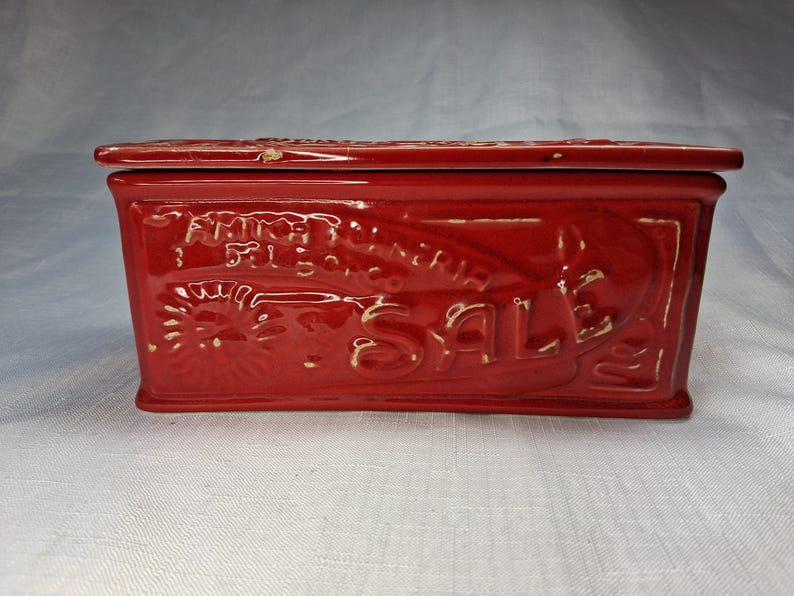 Puede incluir: Una caja rectangular de cer&aacute;mica roja con tapa. La caja presenta texto y dise&ntilde;os en relieve, incluyendo la palabra "SALE" y otro texto ilegible. La caja es probablemente vintage o antigua, con un acabado brillante.