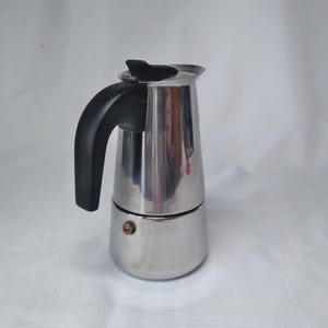 May include: A white ceramic mug with a black handle and the text "But First, Coffee" in a bold, black font. The mug is set against a blurred background, suggesting a cozy setting.