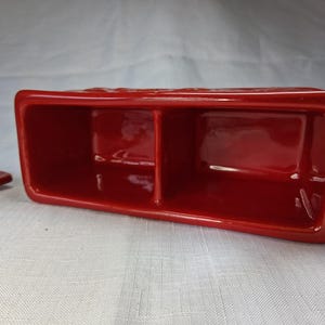 Puede incluir: Un recipiente rectangular de cer&aacute;mica roja con dos compartimentos y una tapa a juego. El recipiente tiene un acabado brillante y probablemente sirve para guardar objetos peque&ntilde;os. La tapa est&aacute; a un lado.