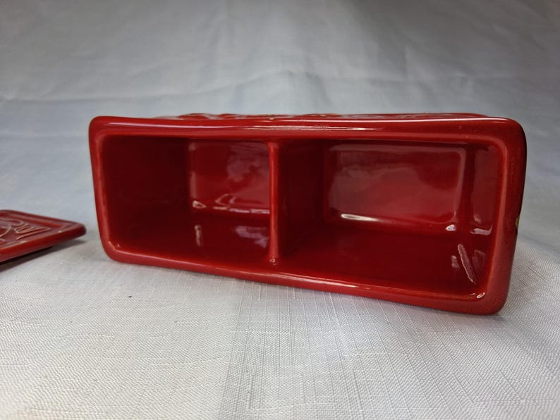 Puede incluir: Un recipiente rectangular de cer&aacute;mica roja con dos compartimentos y una tapa a juego. El recipiente tiene un acabado brillante y probablemente sirve para guardar objetos peque&ntilde;os. La tapa est&aacute; a un lado.