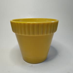 Op de afbeelding: Een gele bloempot met een geribbeld ontwerp rond de rand. De pot heeft een licht taps toelopende vorm en een gladde, glanzende afwerking. De pot is geschikt voor het planten van bloemen of kruiden.