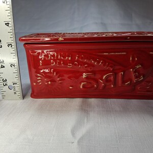 Puede incluir: Una caja rectangular de cer&aacute;mica roja con tapa. La caja presenta texto y dise&ntilde;os en relieve, incluida la palabra "SALE". La caja mide aproximadamente 13 cm de largo.