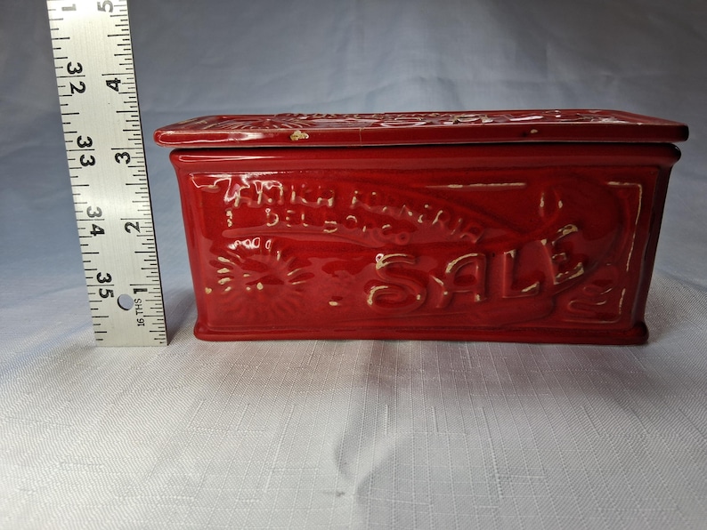 Puede incluir: Una caja rectangular de cer&aacute;mica roja con tapa. La caja presenta texto y dise&ntilde;os en relieve, incluida la palabra "SALE". La caja mide aproximadamente 13 cm de largo.