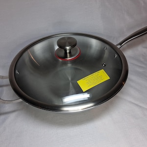 May include: Stainless steel wok with a glass lid. The wok has a long handle and a loop handle on the opposite side. The lid has a stainless steel knob with a red ring. A yellow sticker on the lid reads "TEMPERED GLASS LID CARE".