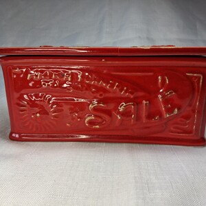 Puede incluir: Caja rectangular de cer&aacute;mica roja con tapa. La caja presenta texto y dise&ntilde;os en relieve, incluida la palabra "SALE". La caja tiene un acabado brillante y parece vintage.