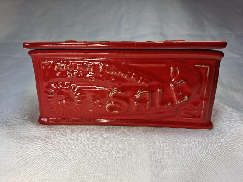 Puede incluir: Caja rectangular de cer&aacute;mica roja con tapa. La caja presenta texto y dise&ntilde;os en relieve, incluida la palabra "SALE". La caja tiene un acabado brillante y parece vintage.