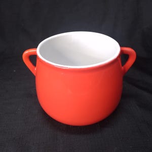 Pode incluir: Uma tigela de cerâmica vermelha vibrante com duas pequenas alças. O interior é branco, contrastando com o acabamento brilhante do exterior. Esta tigela é adequada para servir sopas, ensopados ou outros pratos.