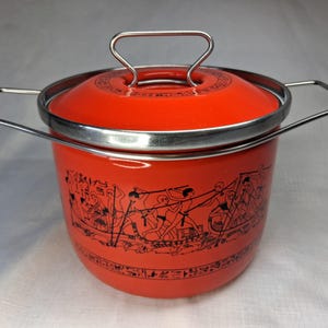 Puede incluir: Una olla vintage de esmalte naranja con tapa y asa y borde de metal plateado. La olla presenta una ilustración negra de figuras en un barco. La olla probablemente se utiliza para cocinar.