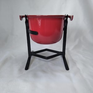 May include: A red enamel bowl with a black metal stand. The bowl has a rounded shape and is supported by a black metal frame with three legs. The stand has a decorative geometric design.