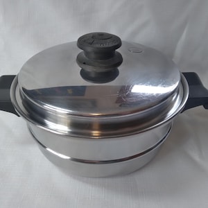 Könnte beinhalten: Edelstahl-Dampfkochtopf mit Deckel und schwarzen Griffen. Der Topf hat eine glänzende, reflektierende Oberfläche und einen schwarzen Knopf auf dem Deckel. Der Topf ist zum Dämpfen von Speisen konzipiert und hat ein mehrschichtiges Design.