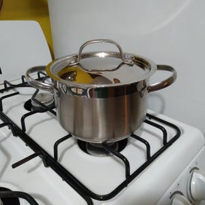 May include: A stainless steel cooking pot with a lid and two side handles sits on a white gas stove with black grates. The pot is reflective, and the stove has a control knob.