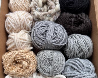 May include: A box filled with various yarn balls in neutral colors. The assortment includes shades of cream, gray, brown, and black. Some yarn is tightly wound, while others have a textured, bulky appearance. The box is made of cardboard.