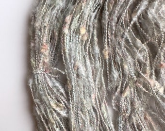 May include: Close-up of a bundle of textured yarn in shades of gray, white, and pastel pink. The yarn has a thick, irregular texture with visible strands and small, colorful flecks. The fibers are loosely woven, creating an airy appearance.