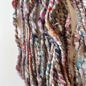 May include: A collection of hand-spun art yarn strands featuring a thick and thin texture. The yarn displays a variegated color palette including shades of pink, blue, green, orange, red, and brown, twisted together with varying fiber densities.