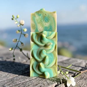 May include: A stack of four handmade soaps with a swirled design in shades of green and tan. The top soap has a carved design. The soaps are displayed on a weathered wooden surface with white flowers and a blurred ocean background.