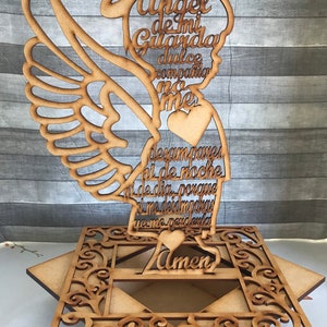 Puede incluir: Una figura de &aacute;ngel de madera con halo y alas. El &aacute;ngel sostiene un coraz&oacute;n y tiene un mensaje escrito en &eacute;l que dice: "Angel de mi Guarda dulce companio no me desampares ni de noche ni de dia porque si me desmayas yo me perderia Amen".