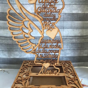Puede incluir: Corte de madera de un &aacute;ngel con alas, halo y coraz&oacute;n. El &aacute;ngel sostiene un rollo con el texto "Angel de mi Guarda dulce compania no me desampares ni de noche ni de dia porque si me desamparas yo me perderia Amen".