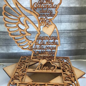 Puede incluir: Figurita de &aacute;ngel de madera con un coraz&oacute;n y alas. El &aacute;ngel sostiene un letrero con el texto "Angel de mi Guarda dulce compania no me desampares ni de noche ni de dia porque si me desamparas yo me perderia Amen".