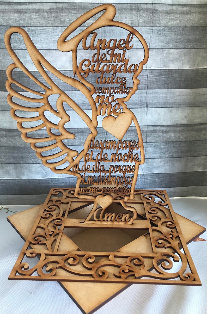 Puede incluir: Figurita de &aacute;ngel de madera con un coraz&oacute;n y alas. El &aacute;ngel sostiene un letrero con el texto "Angel de mi Guarda dulce compania no me desampares ni de noche ni de dia porque si me desamparas yo me perderia Amen".