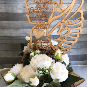 May include: A wooden angel-shaped cake topper with the Spanish text "Angel de mi Guarda dulce compania no me desampares ni de noche ni de dia porque porque mar". The topper is adorned with white flowers and green leaves.