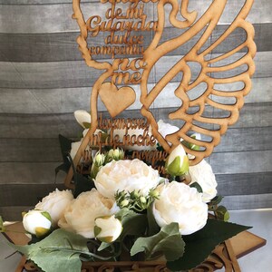 May include: A wooden angel-shaped decoration with the words "Angel de mi Guarda" and other text, placed above white flowers and greenery. The angel has a halo, wings, and a heart. The arrangement sits on a decorative wooden base.