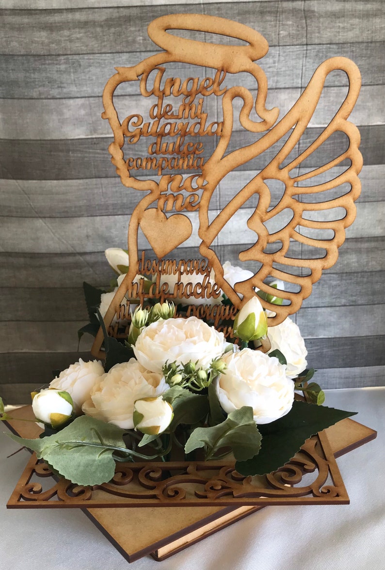 May include: A wooden angel-shaped decoration with the words "Angel de mi Guarda" and other text, placed above white flowers and greenery. The angel has a halo, wings, and a heart. The arrangement sits on a decorative wooden base.