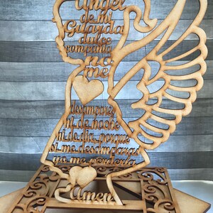 May include: Wooden angel-shaped decoration with cutout wings and a halo. The angel features Spanish text and heart details. The base has an ornate, geometric design. The text includes the word "Amen".