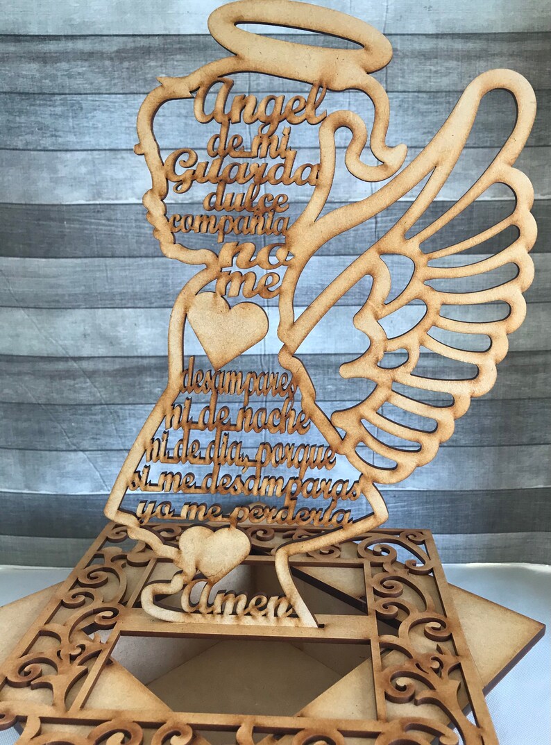 May include: Wooden angel-shaped decoration with cutout wings and a halo. The angel features Spanish text and heart details. The base has an ornate, geometric design. The text includes the word "Amen".