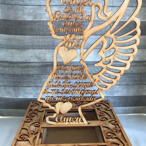 May include: Wooden angel-shaped decorative piece with an openwork design. The angel has a halo, wings, and holds a heart. Text in Spanish is cut into the wood. The base has an ornate, openwork design.
