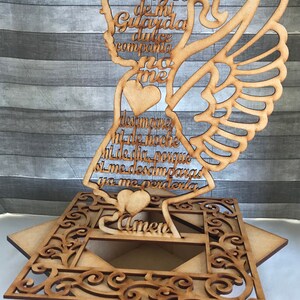 May include: A wooden angel-shaped decoration with an openwork design. The angel has wings, a halo, and text in Spanish. The base is a decorative square with an open center and ornate cutouts. The overall color is light brown.