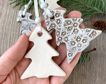 May include: Hand holding several ceramic Christmas tree ornaments. The ornaments are in various shades of white and brown, with textured and smooth surfaces. Each tree has a ribbon for hanging. The background includes pine needles and a wooden surface.