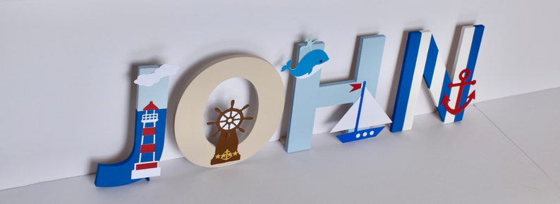 Puede incluir: Letras de madera que deletrean el nombre "JOHN" en azul, blanco y beige. Cada letra tiene un tema n&aacute;utico con un faro, un tim&oacute;n, una ballena, un velero y un ancla.