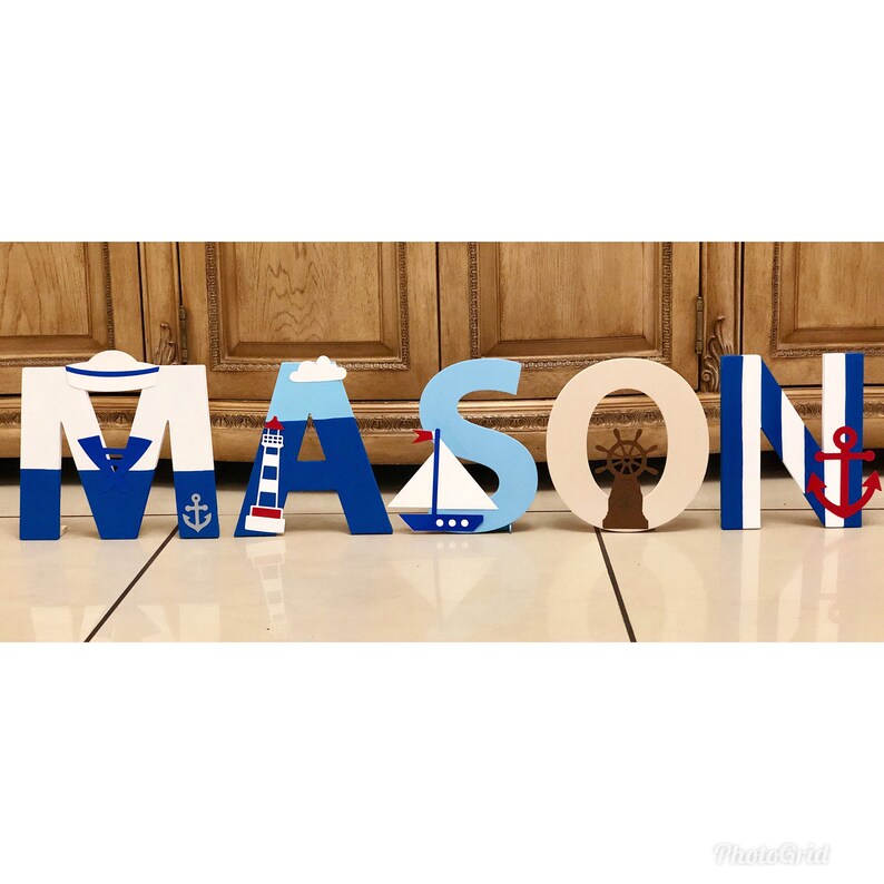 Puede incluir: Letras de madera que deletrean el nombre "MASON" en azul y blanco con decoraciones de tem&aacute;tica n&aacute;utica. La "M" tiene un sombrero de marinero, la "A" tiene un faro, la "S" tiene un velero, la "O" tiene un tim&oacute;n de barco y la "N" tiene un ancla.