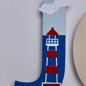 Puede incluir: Una letra J de madera azul con un faro blanco y rayas rojas, una nube blanca y un cielo azul. La letra est&aacute; de pie sobre una superficie blanca.