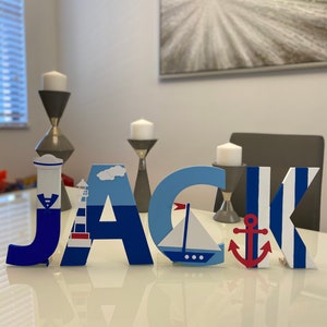 Puede incluir: Letras de madera que deletrean el nombre "JACK" en azul, blanco y rojo. Cada letra presenta un tema n&aacute;utico con un faro, un velero, un ancla y rayas.