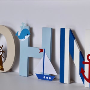 Puede incluir: Letras de madera que deletrean el nombre "JOHN" en azul, blanco y beige. Cada letra tiene un tema n&aacute;utico con un faro, un tim&oacute;n, una ballena, un velero y un ancla.