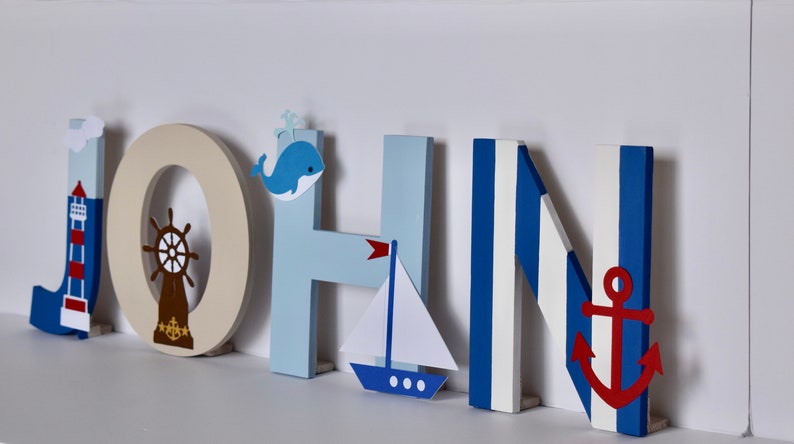 Puede incluir: Letras de madera que deletrean el nombre "JOHN" en azul, blanco y beige. Cada letra tiene un tema n&aacute;utico con un faro, un tim&oacute;n, una ballena, un velero y un ancla.