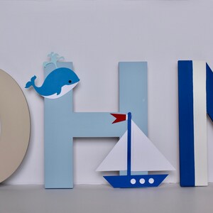 Puede incluir: Letras de madera que deletrean "JOHN" en azul y blanco con decoraciones de tem&aacute;tica n&aacute;utica. La "O" tiene un tim&oacute;n de barco marr&oacute;n, la "H" tiene una ballena azul y la "N" tiene un ancla roja.