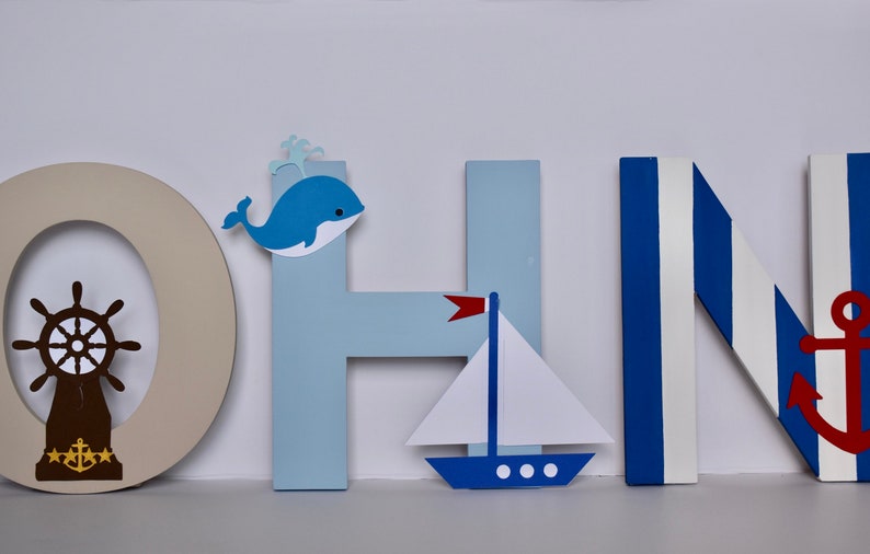 Puede incluir: Letras de madera que deletrean "JOHN" en azul y blanco con decoraciones de tem&aacute;tica n&aacute;utica. La "O" tiene un tim&oacute;n de barco marr&oacute;n, la "H" tiene una ballena azul y la "N" tiene un ancla roja.
