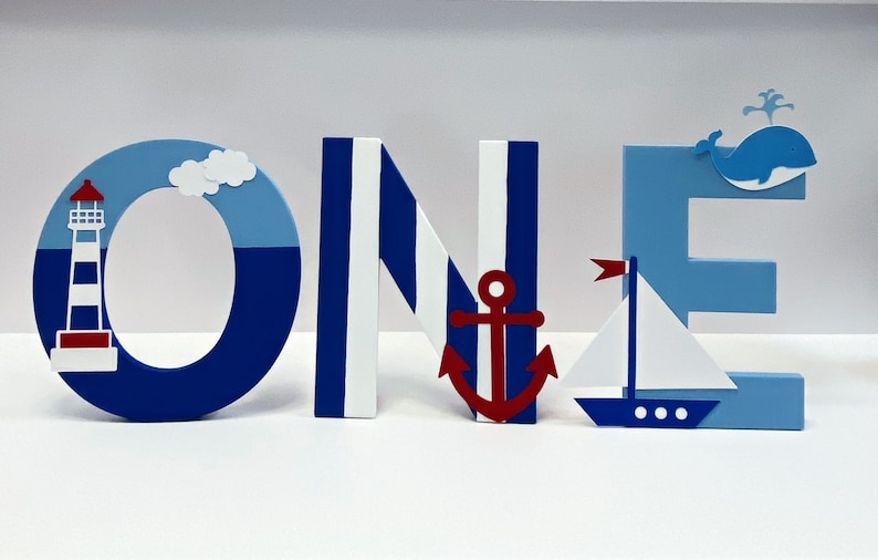 Puede incluir: Letras de madera azules, blancas y rojas que deletrean "ONE" con decoraciones de tem&aacute;tica n&aacute;utica. La "O" tiene un faro y una nube, la "N" tiene rayas blancas y azules, la "E" tiene una ballena y un velero.