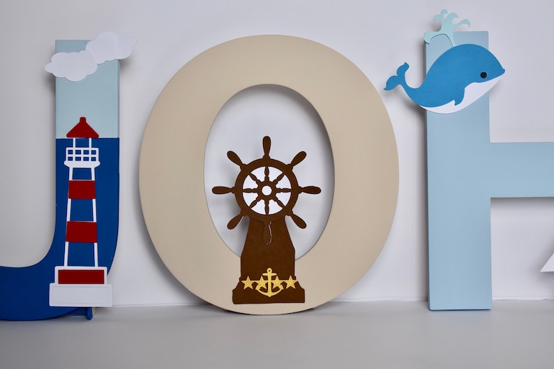 Puede incluir: Letras de madera que deletrean "JOH" en azul, beige y azul claro. La "J" tiene un faro rojo y blanco, la "O" tiene un tim&oacute;n de barco marr&oacute;n con un ancla dorada, y la "H" tiene una ballena azul con vientre blanco.