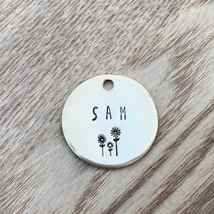 May include: A round, brass dog tag with the name "SAM" engraved on it. The tag also features a design of three stylized flowers.