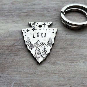 Puede incluir: Chapa de identificación para mascotas de plata en forma de punta de flecha con una escena de montaña, una luna creciente y estrellas. El nombre "Loki" está grabado en la chapa.