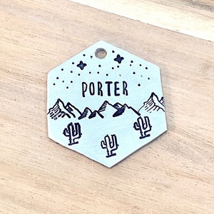 Può includere: Un'etichetta per cani esagonale in argento con un design a catena montuosa nera, tre cactus e il nome "PORTER" inciso su di essa.
