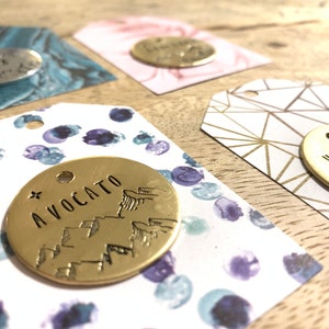 May include: Close-up of several small, round, gold-colored metal tags with various designs and text, including "AVOCATO" and "VANLIFE". The tags are attached to decorative paper tags with patterns in blue, pink, and gold.